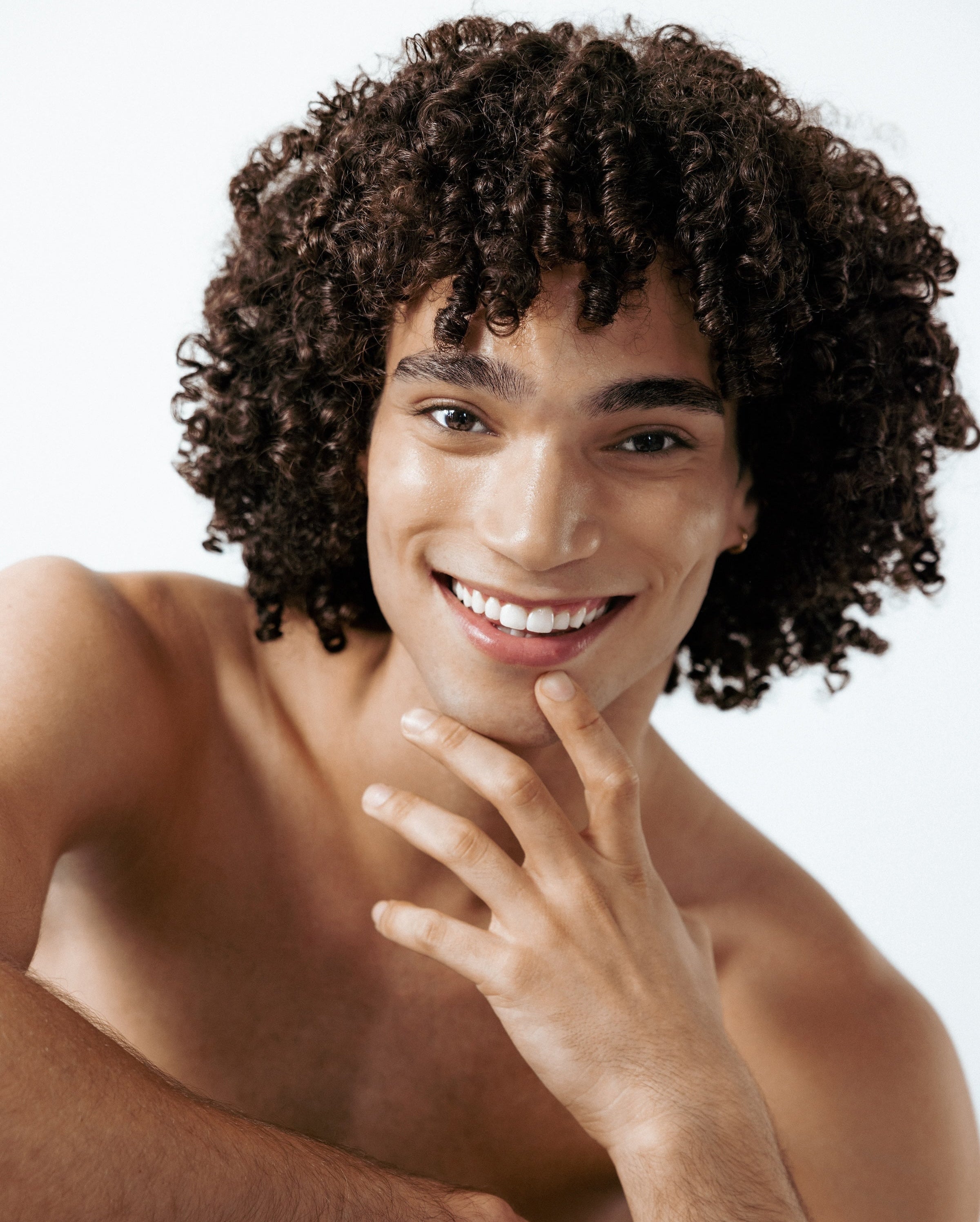 Smiling young man with curly hair touching his clear skin against a white background, representing healthy skin after avoiding harmful DIY acne trends